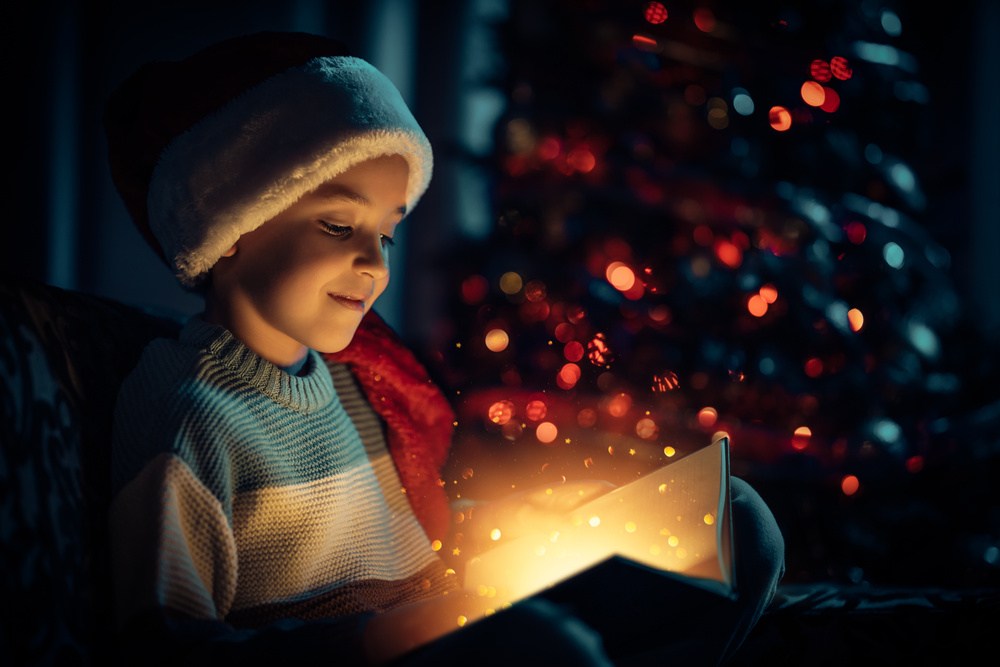 Criança com gorro de Papai Noel lendo um livro natalino em um ambiente alegre e decorado.