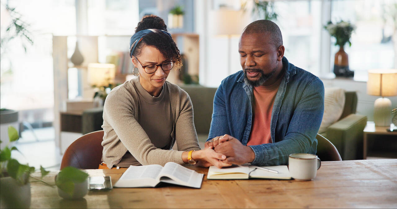 Um casal afro-americano sentado em uma mesa de jantar, estudando a Bíblia e orando. O homem e a mulher estão de mãos dadas, com os olhos fechados em oração. Há uma Bíblia aberta na mesa, junto com um caderno e caneta.
