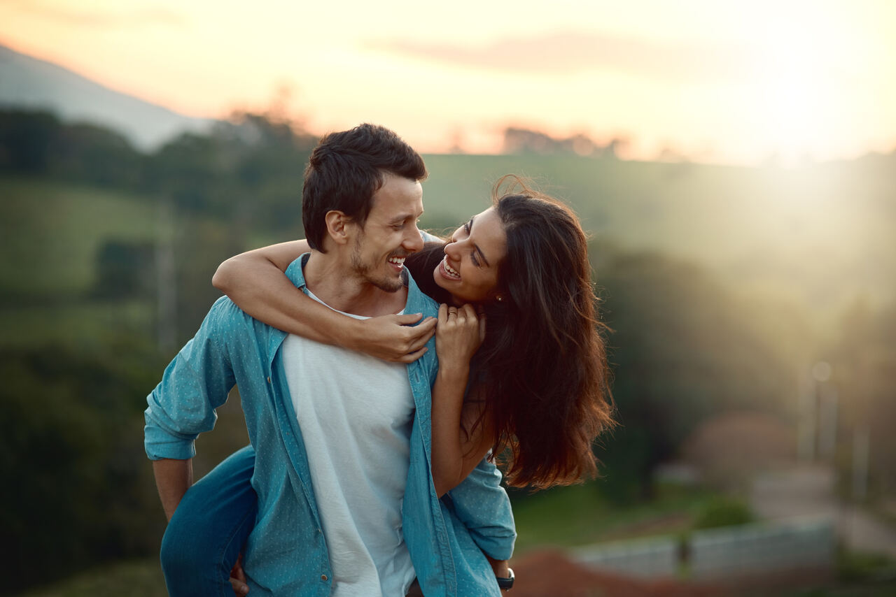 Um casal jovem sorrindo e se abraçando ao ar livre ao pôr do sol. A mulher está sendo carregada nas costas pelo homem, ambos olhando um para o outro com alegria. A mulher está com os braços em volta do pescoço dele. O fundo tem um caminho e vegetação sob uma luz quente.