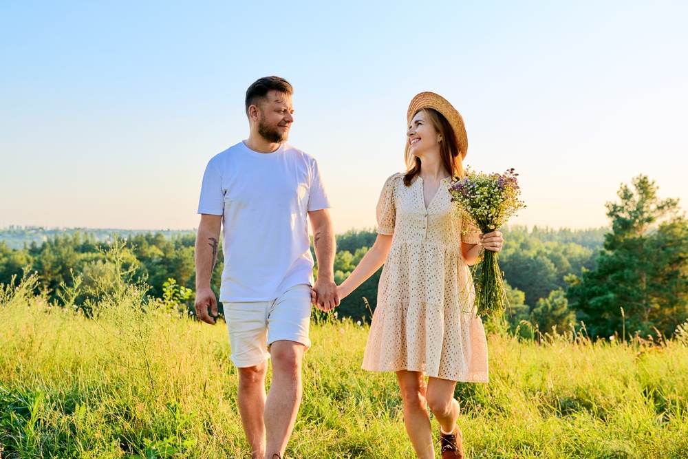 Casal apaixonado caminhando ao ar livre em um campo, mulher com chapéu de palha segurando um buquê de flores, sob luz do pôr do sol.