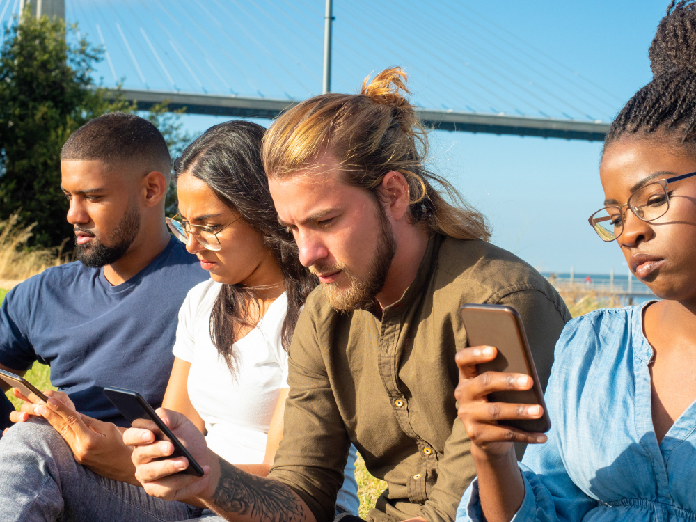 Grupo de jovens usando smartphones ao ar livre, sentados juntos em um parque sob céu azul, na presença de uma ponte na fundo, em um dia ensolarado.